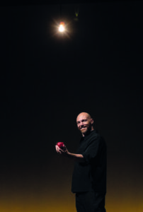 Andrea Pezzi sorridente con una mela rossa in mano, illuminato da una lampadina su sfondo scuro.