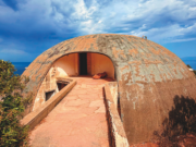 No time no space, il mito della casa che veniva da un altro pianeta. Viaggio nella Cupola abbandonata di Dante Bini a Costa Paradiso, dove Michelangelo Antonioni e Monica Vitti costruirono il loro rifugio impossibile. Struttura a cupola in cemento affacciata sul mare, immersa nella vegetazione mediterranea sotto un cielo nuvoloso.