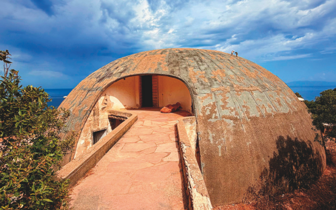 Struttura a cupola in cemento affacciata sul mare, immersa nella vegetazione mediterranea sotto un cielo nuvoloso.