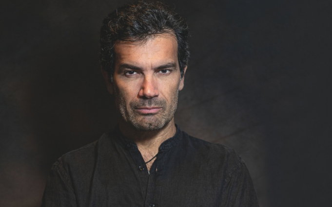 Ritratto di Marco Bonini con camicia nera e braccia incrociate su sfondo scuro.