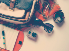 La borsa torna a fare la borsa Accessori da viaggio con borsa, taccuino, occhiali da sole, fotocamera e orologio disposti su un piano.