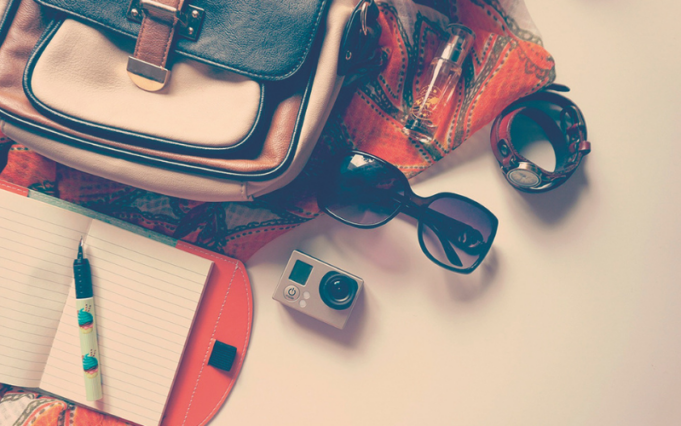 Accessori da viaggio con borsa, taccuino, occhiali da sole, fotocamera e orologio disposti su un piano.
