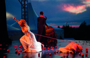 Scena teatrale al tramonto con una donna in abito bianco legata da una corda, attori sul palco e mele rosse sparse a terra.