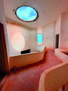 Interno di una struttura a cupola con oculo sul soffitto, pareti curve e vista sul mare.