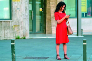 Donna con abito rosso usa lo smartphone in uno spazio urbano, con borsa a tracolla.